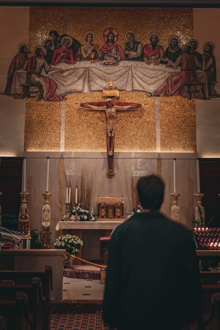 A man stands in prayer before an altar with religious mosaics and cross in a solemn church setting.