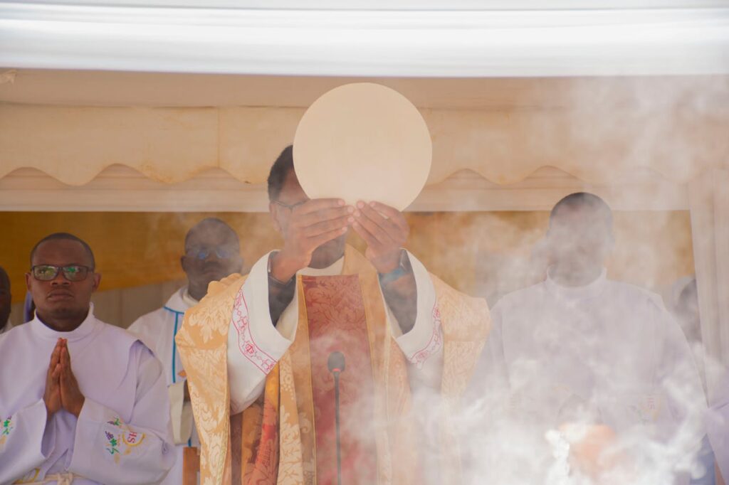 A priest in ceremonial robes holds the Eucharist wafer during a religious service, surrounded by incense smoke.